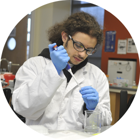 A person in lab coat and gloves holding a glass.